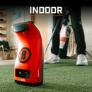 An orange Bushnell golf launch monitor sits on green indoor turf. A golfer stands in the background preparing to swing a club. Text overlay reads "INDOOR."