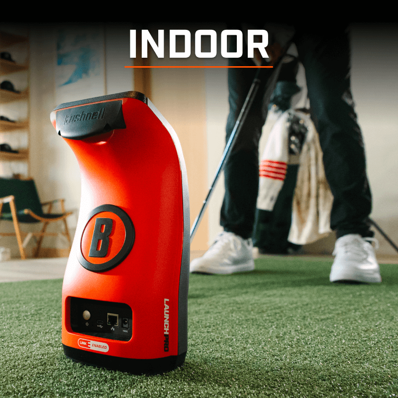 An orange Bushnell golf launch monitor sits on green indoor turf. A golfer stands in the background preparing to swing a club. Text overlay reads "INDOOR."